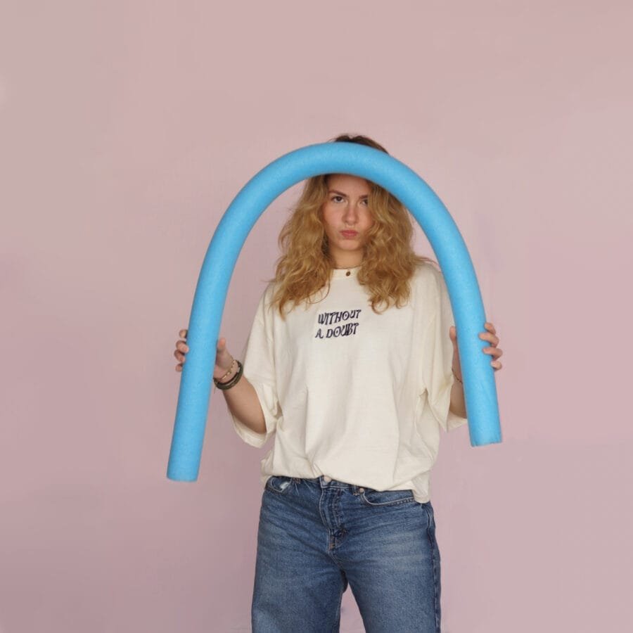 Female model wearing a cream oversized T-shirt with a small front graphic. She is holding a pool noodle.